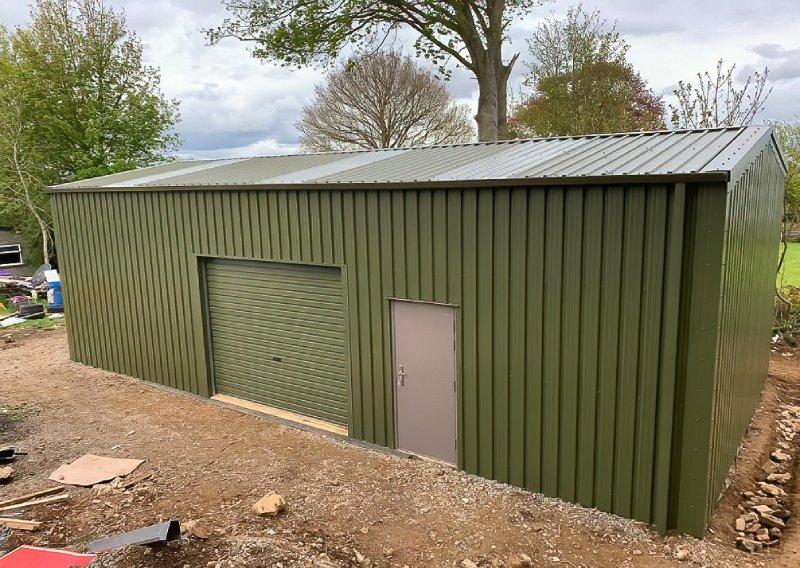 Elevated view of a green steel shed featuring translucent roof panels, a roller door, and a personnel door, surrounded by a dirt construction area.